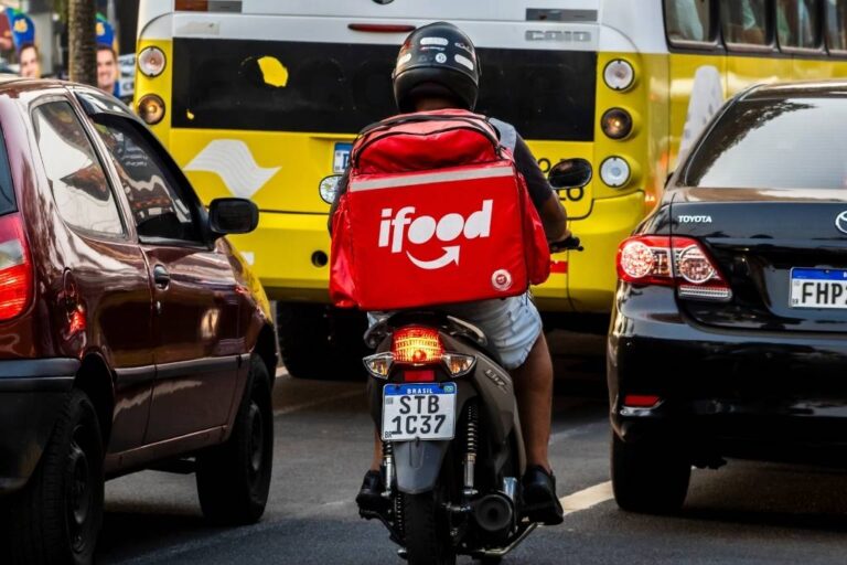 Pessoa entregadora do iFood em moto circulando entre carros e ônibus em via urbana. A imagem representa prudência no trânsito e atenção redobrada nas ruas.